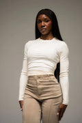 Woman wearing a white long-sleeve top and beige pants against a gray background