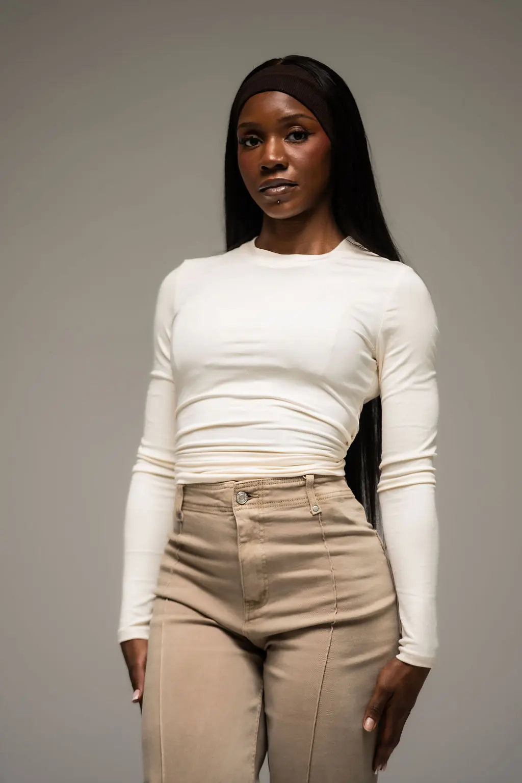 Woman wearing a white long-sleeve top and beige pants against a gray background